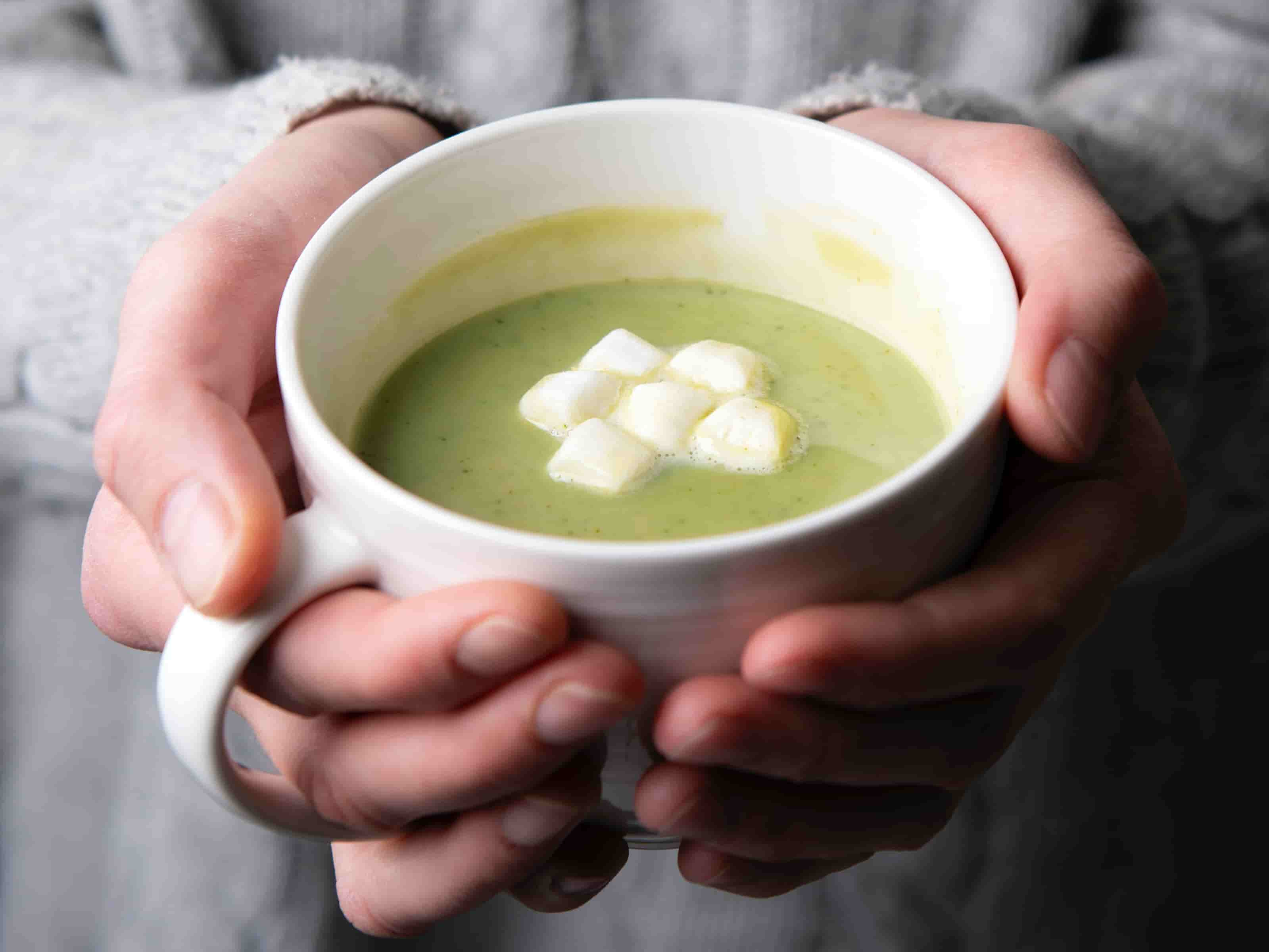 Gruene Fluessigkeit in weißer Tasse mit Marshmallows darauf, gehalten von zwei Haenden