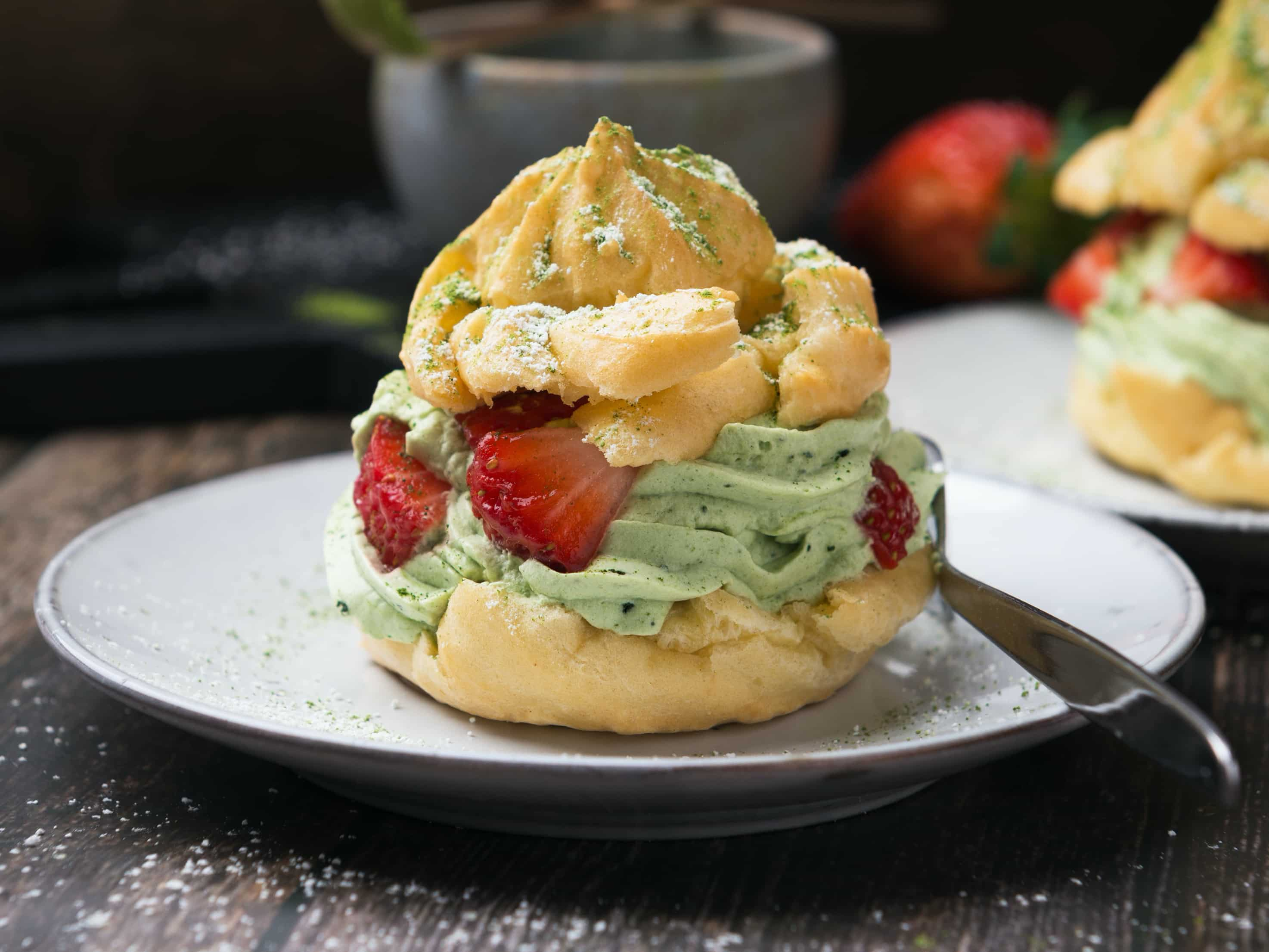 aufgeschnittener Windbeutel, gefuellt mit gruener Matcha Sahne und Erdbeerstuecken, auf Teller mit Gabel daneben